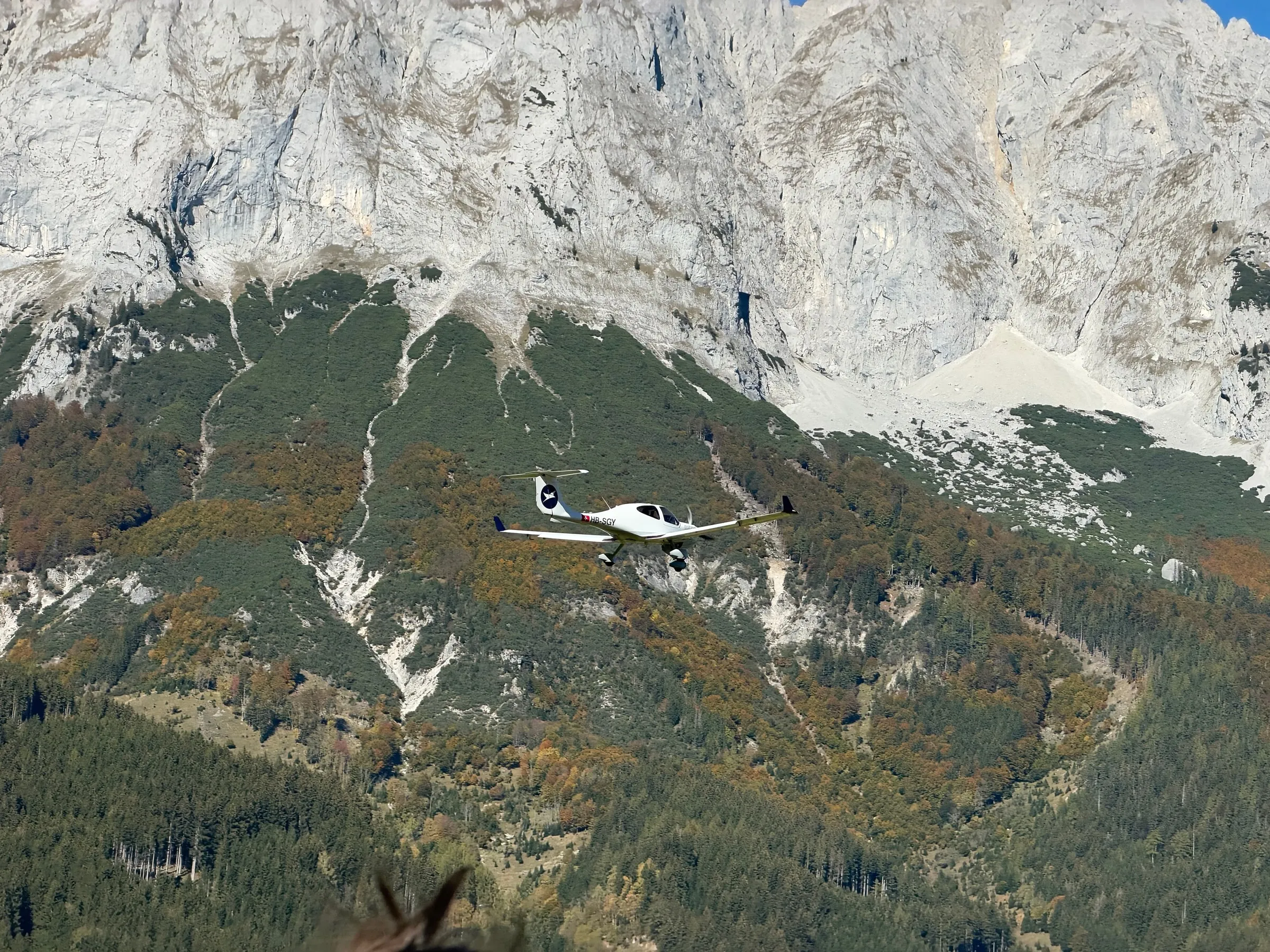 Diamond DA40 HB-SGY in flight against the steep rock face and wooded slopes of the Grimming